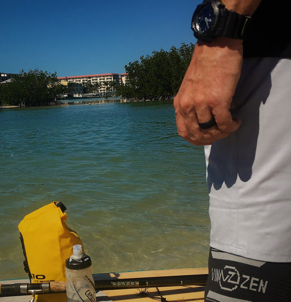 man wearing black silicone rubber ring similar to qalo enso honor outside while paddleboarding in ocean wearing knee sleeve and casio g shock watch